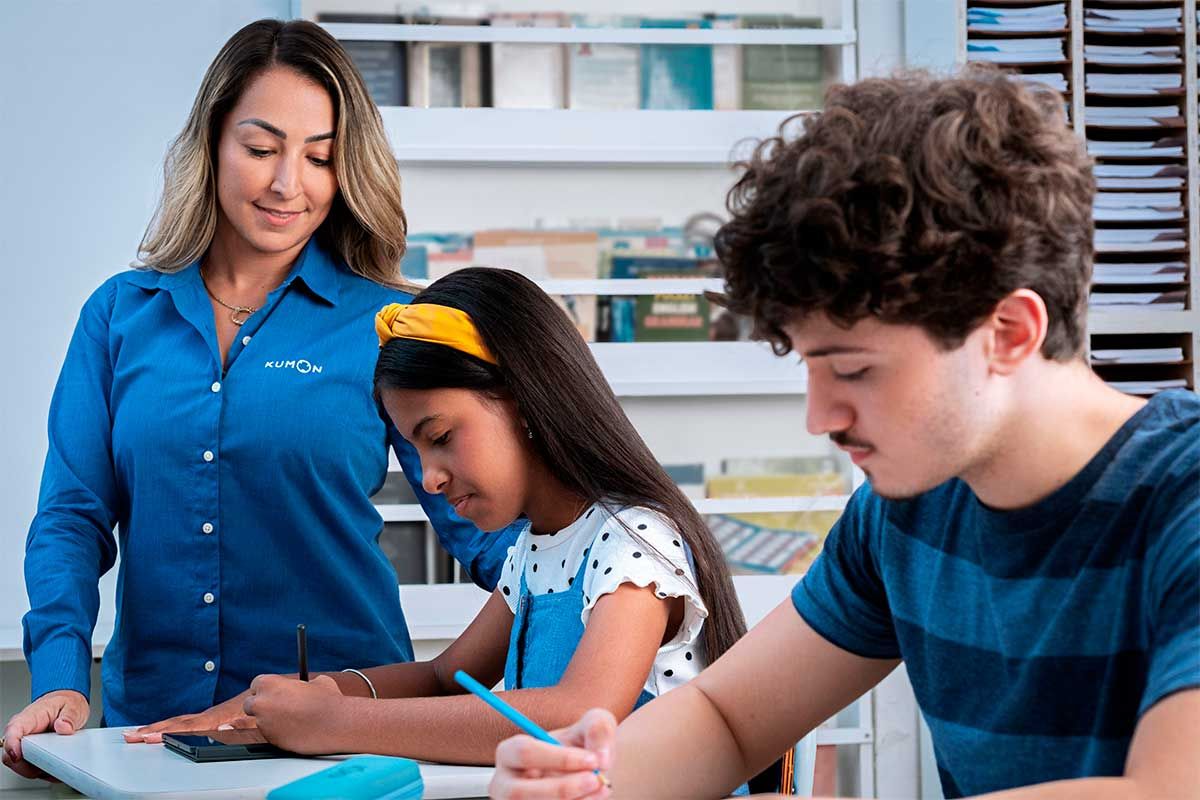 Dois alunos estudando de forma concentrada com a orientadora do método Kumon observando a execução
